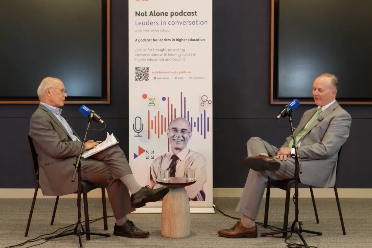 Two men sitting down speaking into microphones with a sign in between them that says "Not Alone podcast" 