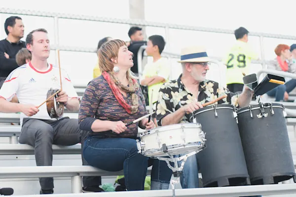 Three supporters in metal bleachers play percussion: a man with a tambourine, a woman on a snare drum, and an older man in a straw hat on tall drums.