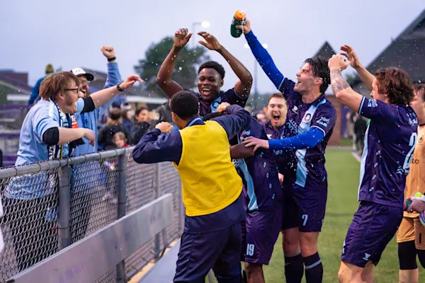 AFC Ann Arbor players in purple kits celebrate at a fence with cheering Main Street Hooligans fans in light blue as one player sprays a water bottle.