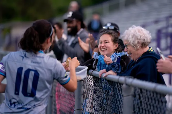 Smiling supporter in a blue scarf offers cookies over a chain-link fence to an AFC Ann Arbor player in a No. 10 jersey as fans laugh and clap.