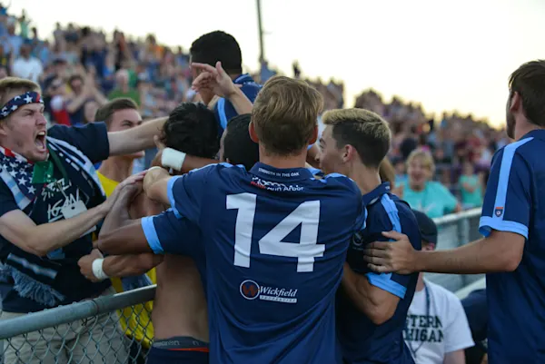 AFC Ann Arbor players in navy jerseys pile together at a fence with a shouting fan in a stars-and-stripes bandana as packed stands look on.