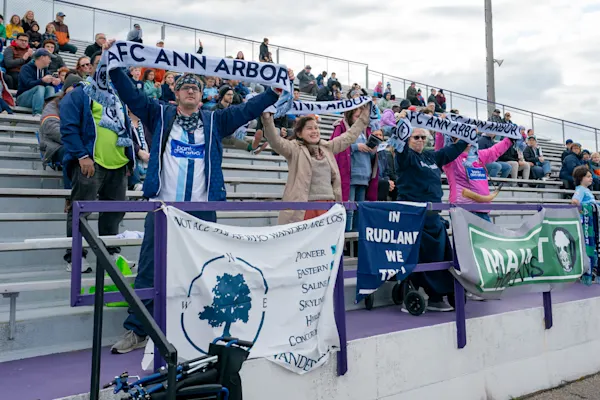 Main Street Hooligans fans hold AFC Ann Arbor scarves overhead in the bleachers, with supporter banners hung on a purple railing below.