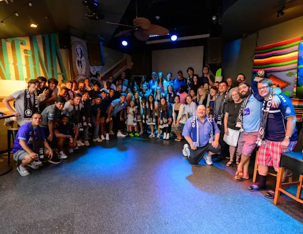 Supporters, staff, and players in navy and white scarves pose as a large group in a dim, blue-lit bar with a mural and striped blanket on the walls.