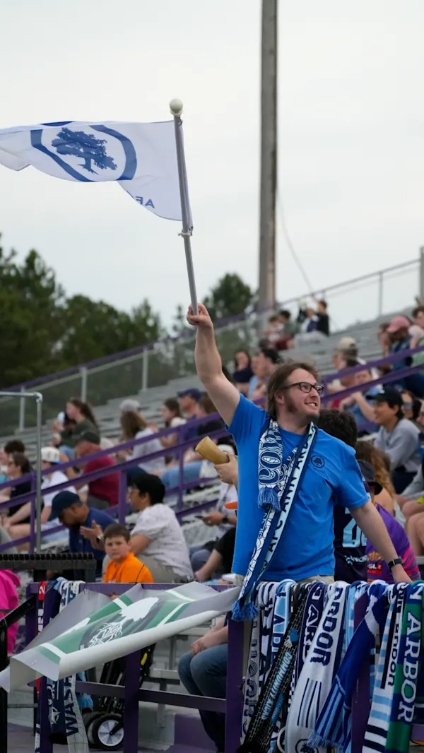 A smiling Main Street Hooligans fan in a blue shirt and scarf waves a white AFC Ann Arbor tree flag in the stands above a railing draped with scarves.
