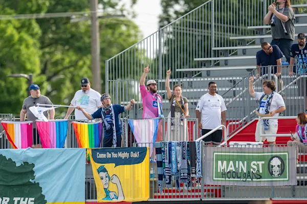Main Street Hooligans fans in metal bleachers raise one finger above pride flags, hanging scarves, and a Main St Hooligans banner.