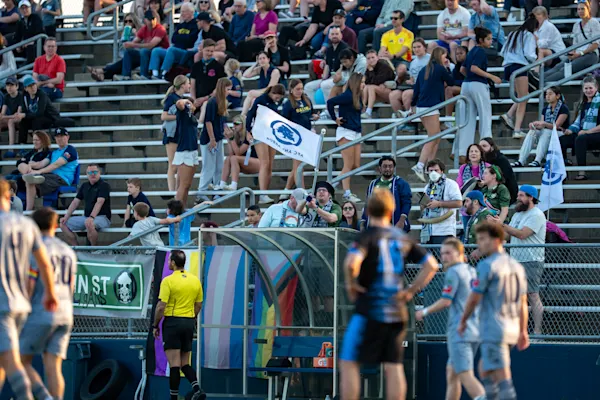 Main Street Hooligans fans wave AFC Ann Arbor flags in the bleachers above Pride flags and a supporters' banner, with blurred players on the field.