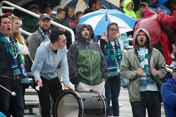 Main Street Hooligans fans in green and blue scarves sing in the rain on metal bleachers as one plays a bass drum, with umbrellas and ponchos behind.