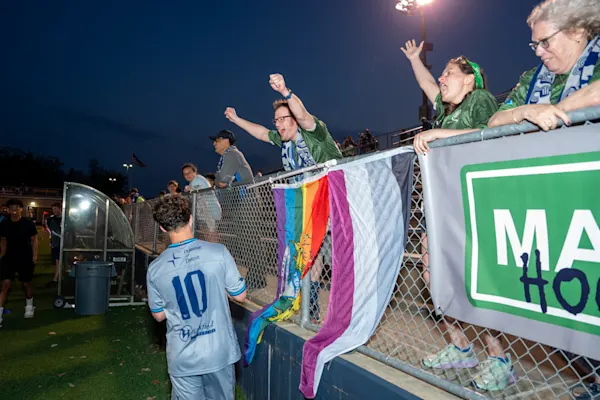 Main Street Hooligans fans in green jerseys and scarves cheer at a fence draped with pride flags as a player in a No. 10 jersey faces them at night.