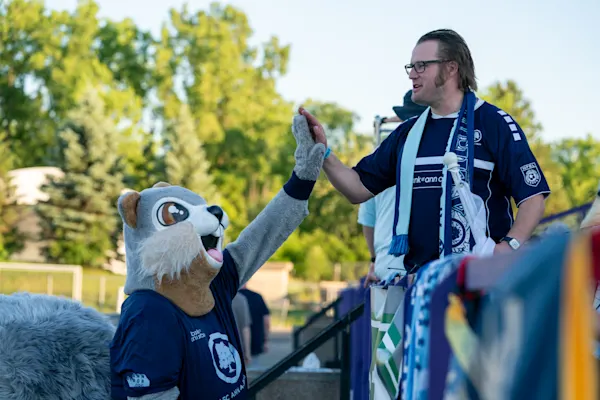 A Main Street Hooligans fan in a navy jersey and scarf high-fives Nutmeg, the squirrel mascot of AFC Ann Arbor, beside scarves draped on a railing.