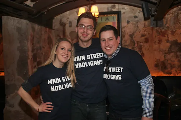 Three smiling fans in navy "Main Street Hooligans" T-shirts pose arm in arm in a dimly lit bar with rough stone walls and a lamp overhead.