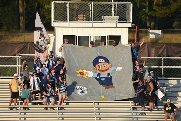 Main Street Hooligans in scarves hold a large painted banner of a mustached cartoon man in an AA cap reaching for a star on metal bleachers.