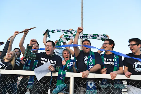 Main Street Hooligans members in navy shirts shout and raise fists and green-and-white Ann Arbor scarves behind a fence as blue streamers fly overhead.