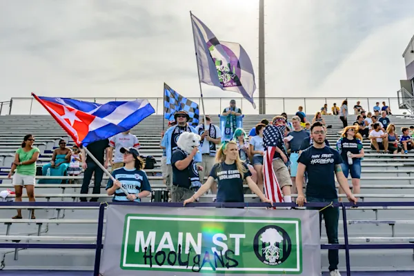 Main Street Hooligans fans wave Cuban, checkered and club flags in stadium bleachers behind a green Main St Hooligans banner, one in a white wolf mask.