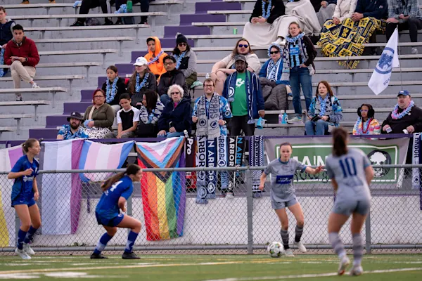 Main Street Hooligans in scarves and blue leis watch from bleachers above pride flags and scarves on a fence as women players contest the ball.