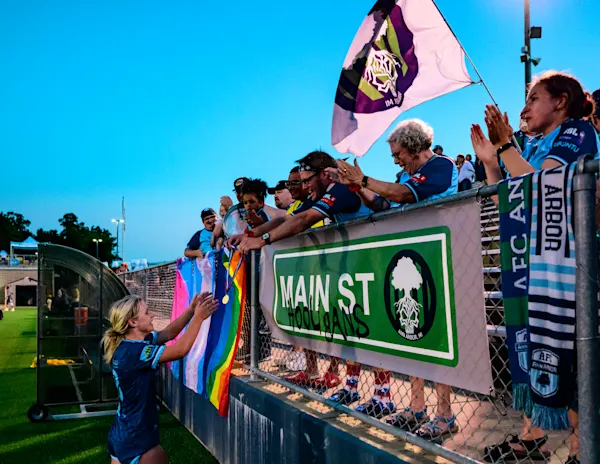 Chloe Ricketts reaches up for a medal from cheering Main Street Hooligans fans at a fence draped with Pride flags, a scarf and the MSH banner at dusk.