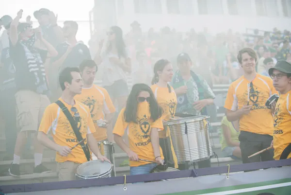 Drummers in yellow "Vencedores" shirts play snare and bass drums amid hazy smoke, with cheering fans in scarves filling the stands behind them.
