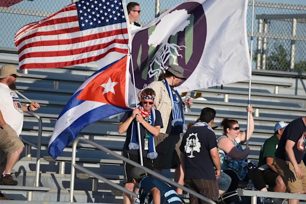 Fans in blue and white scarves wave an American flag, a Cuban flag, and a large white flag with a tree logo in sunny stadium bleachers.