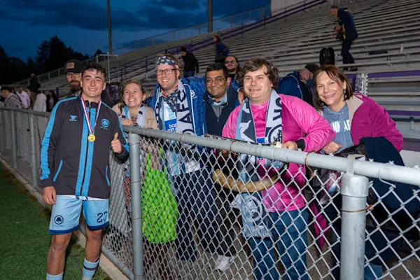 Smiling Main Street Hooligans fans in scarves pose behind a chain-link fence with a medal-wearing AFC Ann Arbor player giving a thumbs-up at dusk.