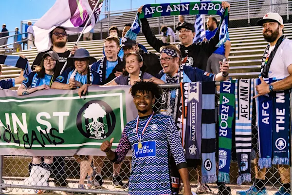 Smiling AFC Ann Arbor player wearing a medal gives a thumbs-up before cheering Main Street Hooligans fans holding scarves, flags, and a banner behind a fence.