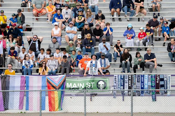 Main Street Hooligans fans cheer from metal bleachers above a fence hung with pride flags, a Main St Hooligans banner, and AFC Ann Arbor scarves.
