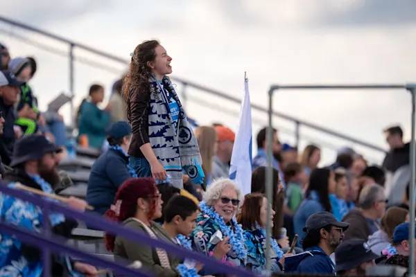 A Main Street Hooligans fan in a gray and navy scarf stands and sings in the bleachers among seated fans wearing blue leis and Hawaiian shirts.