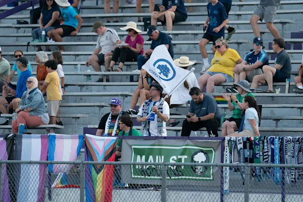 Main Street Hooligans fans in the stands, one waving an AFC Ann Arbor flag, behind Pride flags, scarves, and a Main St Hooligans banner on the fence.