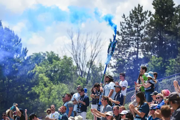 Main Street Hooligans fans in scarves cheer in sunny stands as blue smoke billows from a raised flag, with tall trees behind them.
