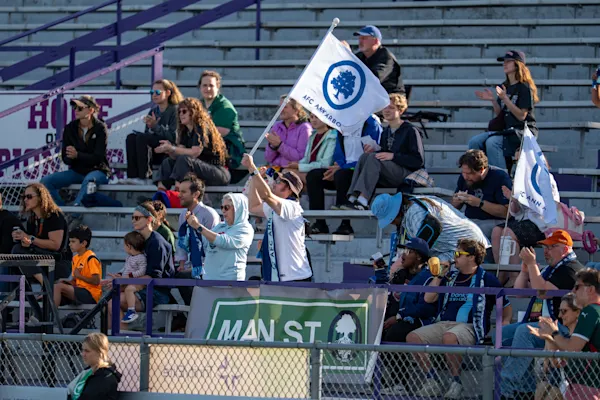 Main Street Hooligans fans in scarves cheer and wave white AFC Ann Arbor tree flags in sunlit bleachers above a green "MAIN ST" banner.