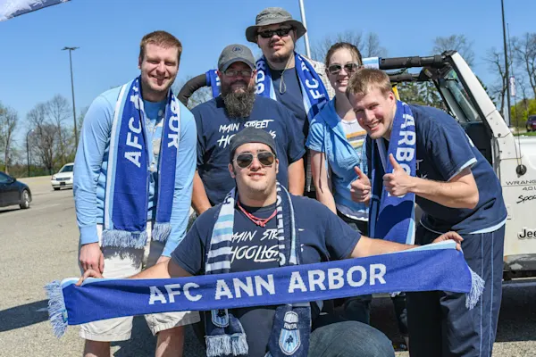 Six Main Street Hooligans fans in blue scarves pose by a white Jeep in a sunny parking lot, one holding up an AFC Ann Arbor scarf, one giving thumbs up.