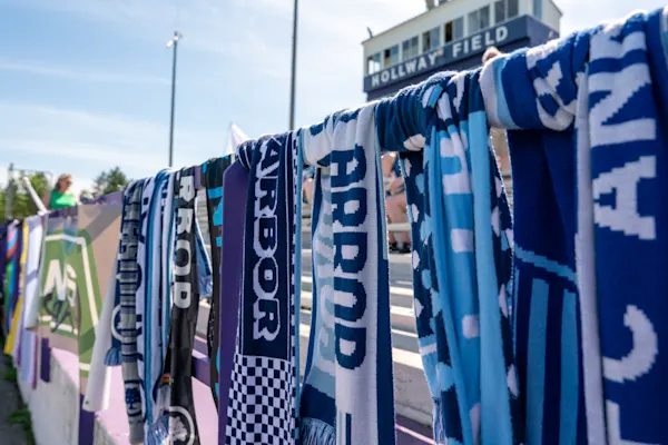 Knit blue and white scarves reading "Arbor" hang along a stadium railing with banners and bleachers beneath the Hollway Field press box.