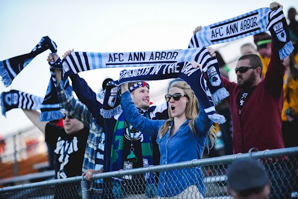 Main Street Hooligans fans hold navy and light blue AFC Ann Arbor scarves overhead and cheer behind a chain-link fence in the stands.