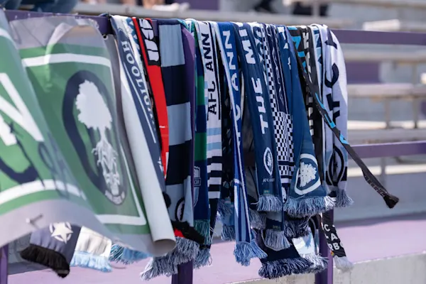 Dozens of blue, white, and black AFC Ann Arbor and Hooligans scarves hang on a purple stadium railing beside a green banner with a tree logo.