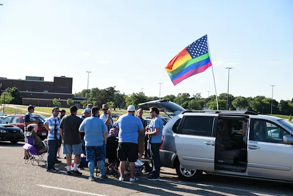 Main Street Hooligans fans in light blue and scarves tailgate by a silver minivan in a parking lot, a rainbow American flag flying above them.