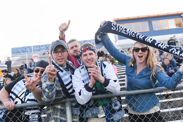 Smiling supporters in striped AFC Ann Arbor scarves cheer at a stadium railing as a woman holds up a "Strength in the Oak" scarf.