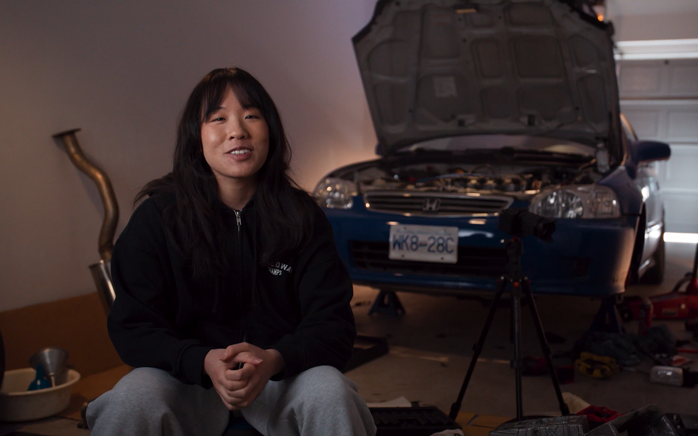 A woman smiling, sitting in front of a car with the hood up