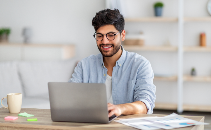 Person wearing glasses using a laptop at a desk, with papers and a coffee cup nearby in a home office setting