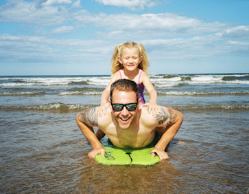 A dad uses a body board in the sea with his young daughter.