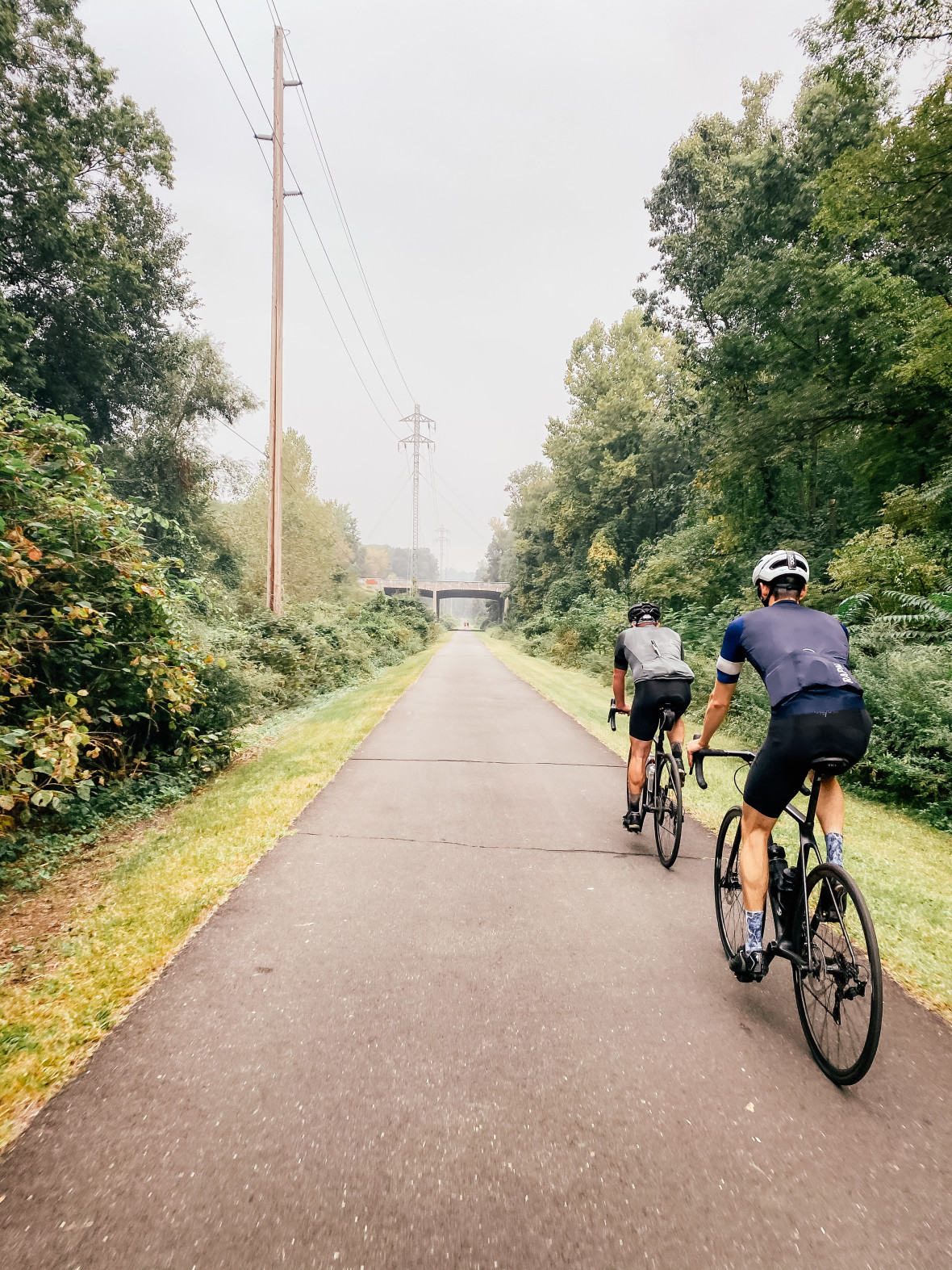 Two bicyclists riding a paved bike path towards bridge underpass in distance