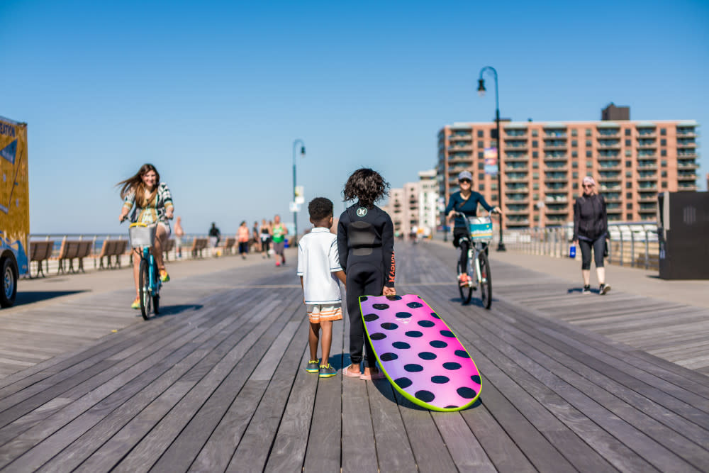 Two children dragging a surfboard down the boardwalk at Long Beach while bikers pass them