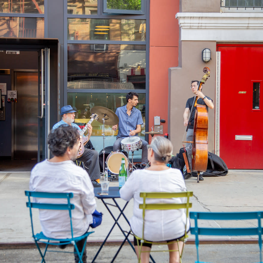 Band plays on sidewalk while two individuals sitting at a table with folding chairs in an open NYC street at Car Free Earth Day 2021