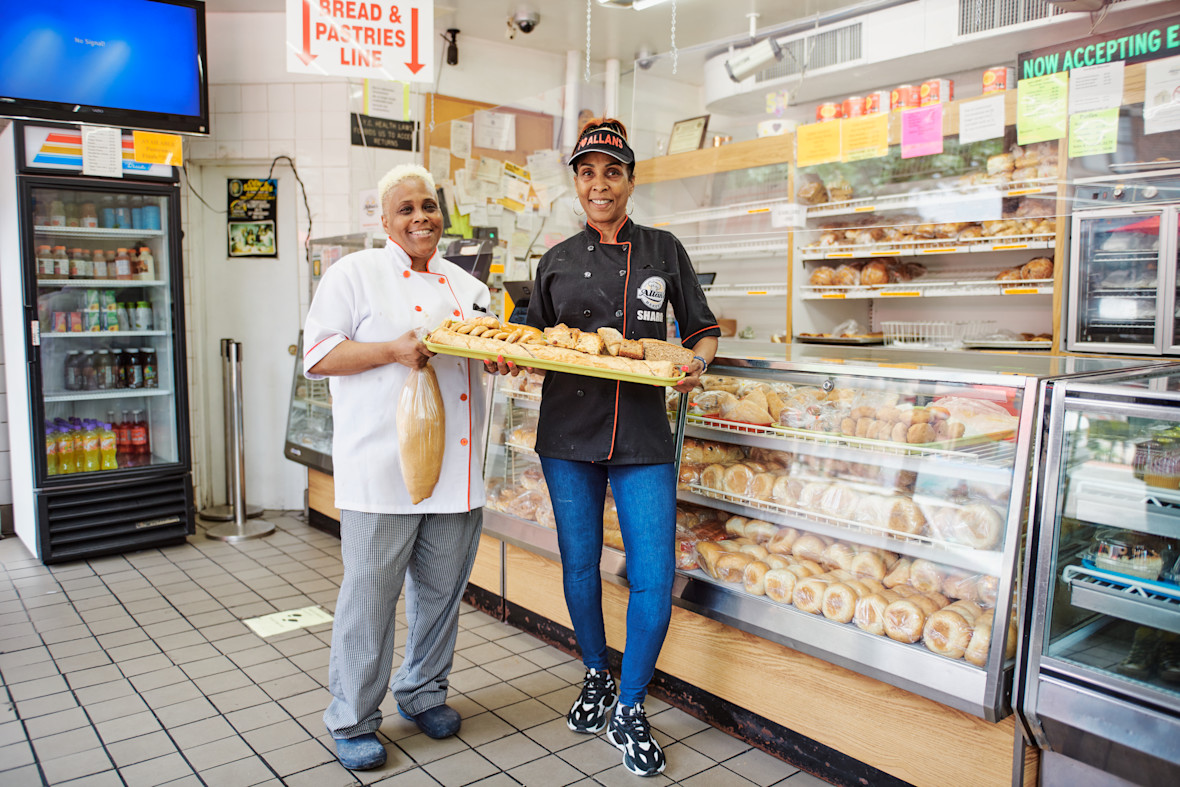 Two Allan's Bakery employees holding a tray of baked goods while standing in front of bakery case