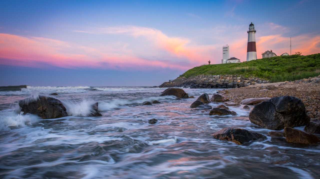 Montauk Lighthouse with the sun setting behind it in pinks and oranges against the blue sky