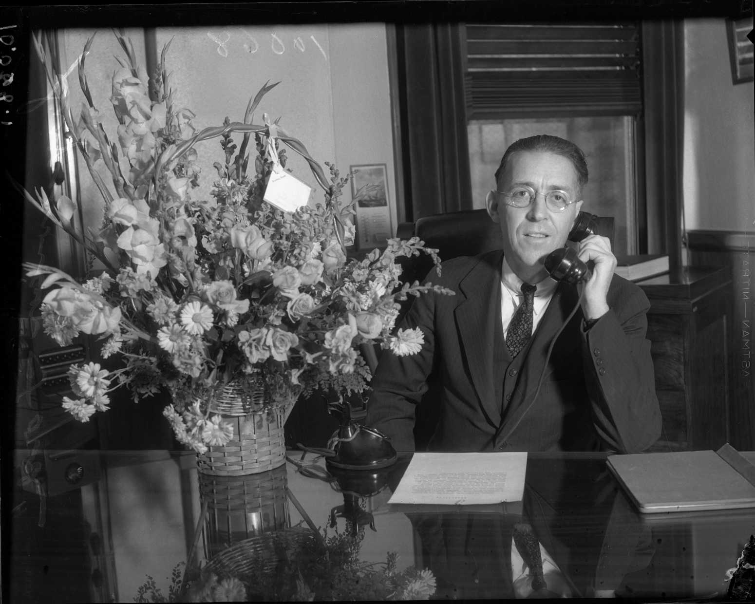 Supervisor John Anson Ford sits at a desk with a bouquet of flowers, 1930s