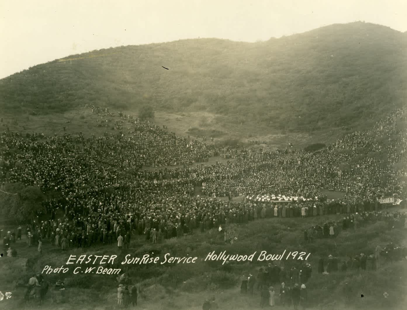 The first Easter Sunrise Service at the Hollywood Bowl drew 10,000 people in 1921. A large crowd at the first Easter Sunrise Service at the Hollywood Bowl, 1921.
The Los Angeles Philharmonic Orchestra sits with white sheet music, and most people are standing or sitting on the ground.