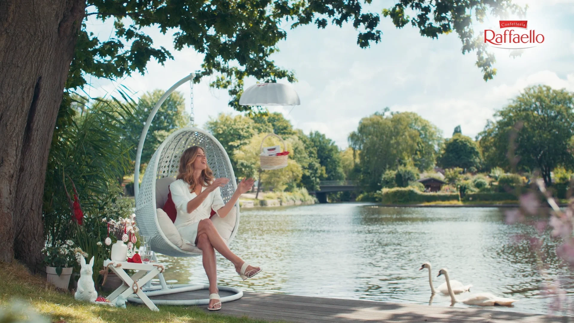 a beautiful woman in a white dress is sitting in an hanging chair at a lake trying to catch the basket from under a white parachute