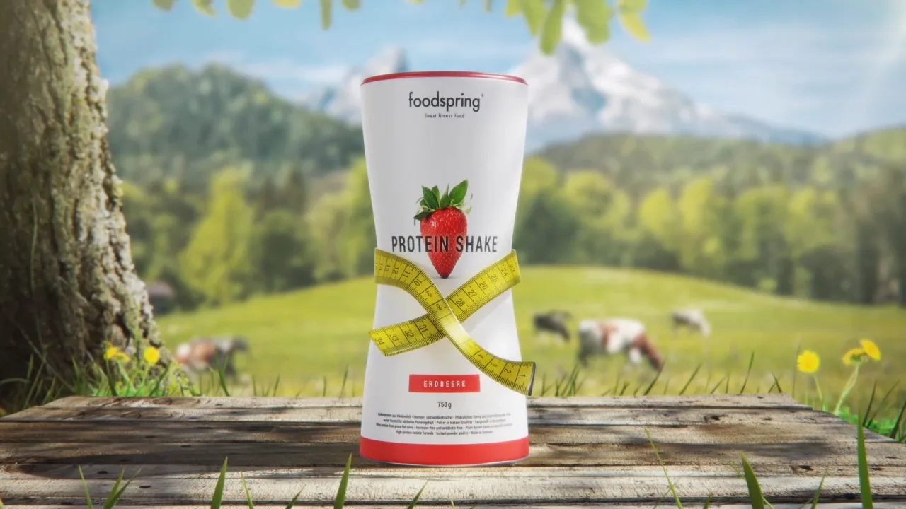 a foodspring strawberry protein shake packshot having a measuring tape around its hips standing on a table in from of a tree and a field with cows on it.