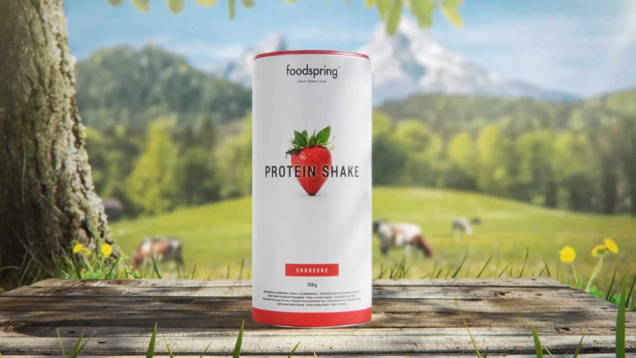 a foodspring strawberry protein shake packshot standing on a table in from of a tree and a field with cows on it.