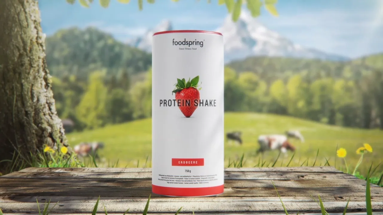 a foodspring strawberry protein shake packshot standing on a table in from of a tree and a field with cows on it.