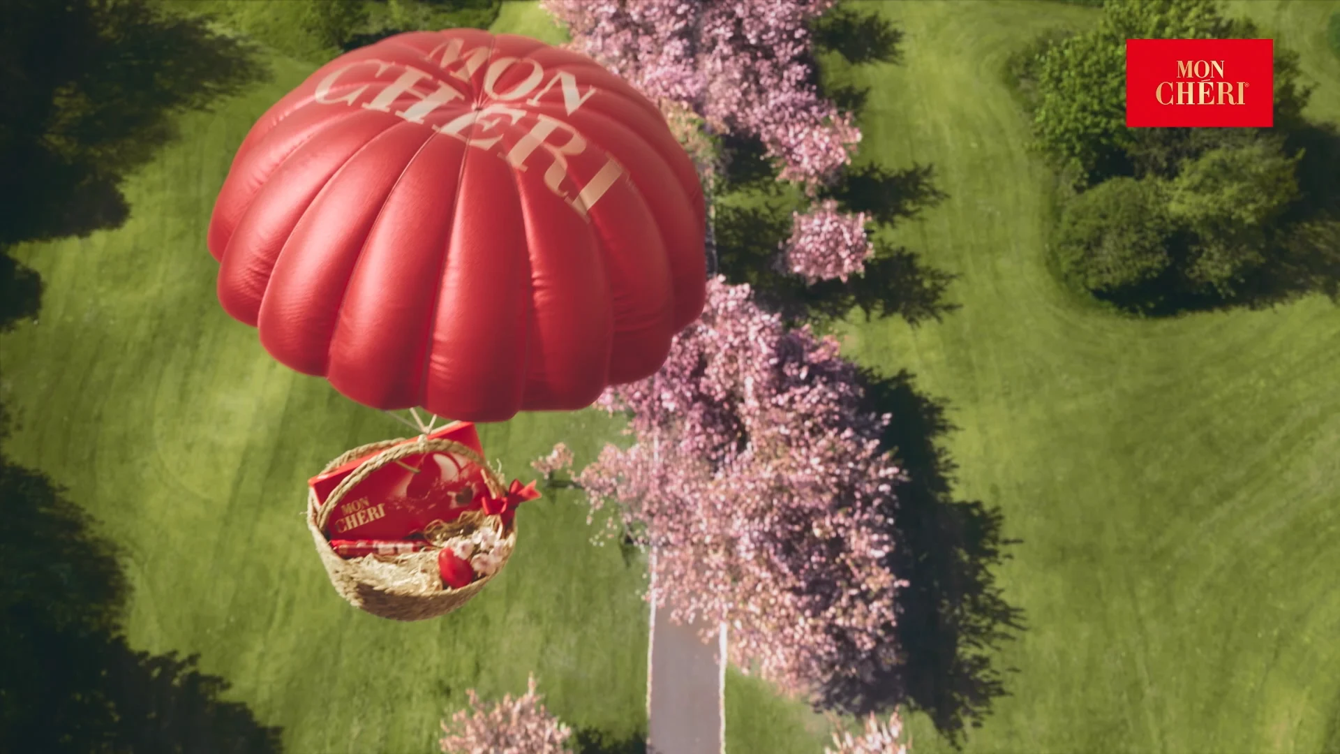 a top shot in which a red Mon Cheri parachute with a basked underneath is flyind over a street and green fields