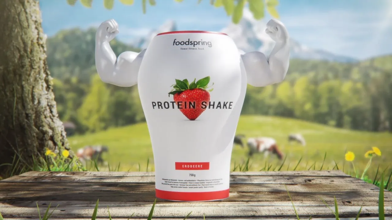 a foodspring strawberry protein shake packshot showing muscles with his comic arms standing on a table in from of a tree and a field with cows on it.